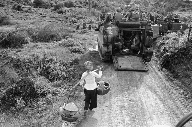A young Vietnamese woman uses a couple of baskets to carry a baby and her shopping as she passes a line of Australian troops resting in an armored personnel carrier 